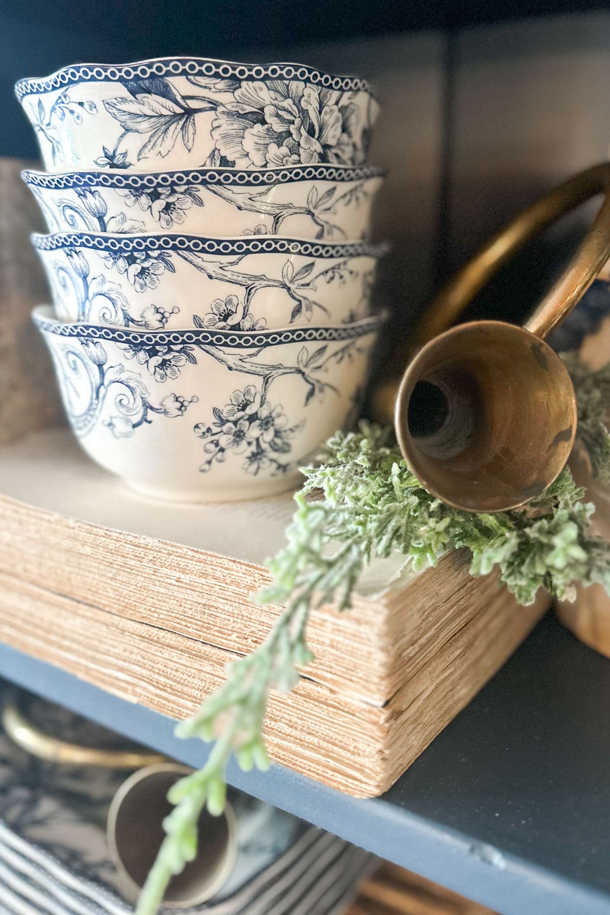 Stack of four white bowls with blue floral patterns, placed on a pile of beige books. A brass horn ornament with green foliage is nearby. The arrangement is on a dark wooden shelf.