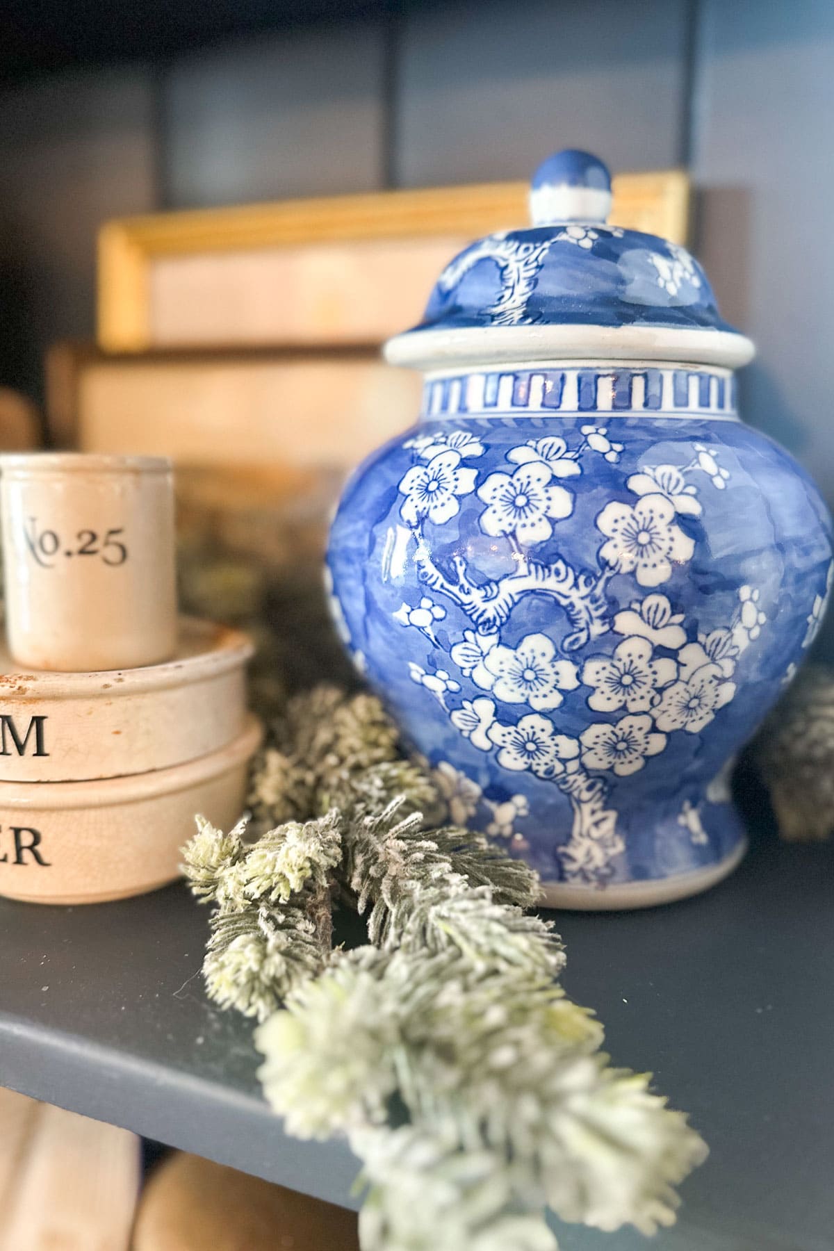 A decorative blue and white floral ceramic jar with a lid is displayed on a shelf next to rustic beige containers and greenery. Framed pictures are blurred in the background.