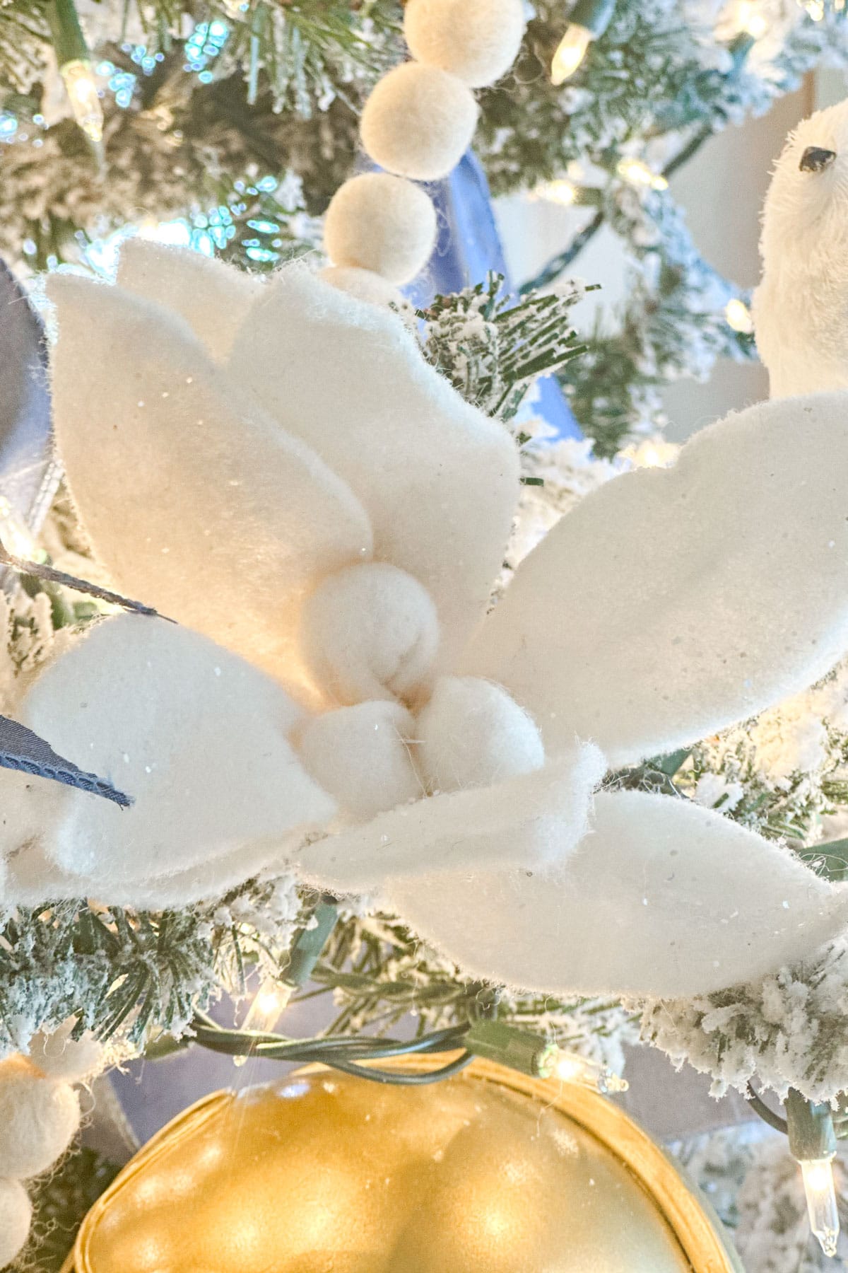 A close-up of a Christmas tree adorned with white, felt magnolia flowers, blue ribbon, and warm twinkling lights. Snow-like textures cover the branches, creating a frosty, elegant winter atmosphere.