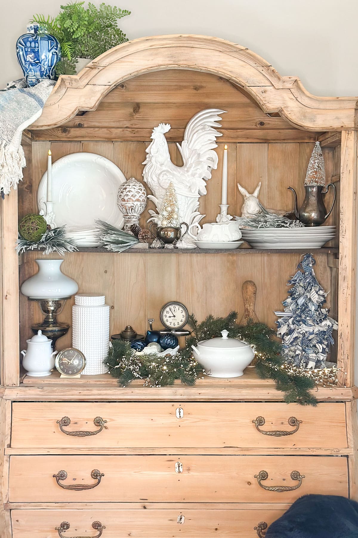 A wooden dresser with a hutch decorated for Christmas. It features ceramic roosters, silver tea sets, greenery, candles, and festive ornaments. A blue vase and a cloth are on top, and clocks and crockery are displayed on the shelves.