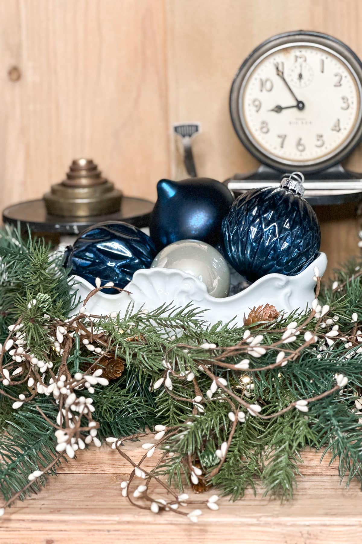 A decorative arrangement featuring green pine branches, white berries, a white ceramic bowl with blue and silver ornaments, and an antique-style clock and lamp in the background on a wooden surface.