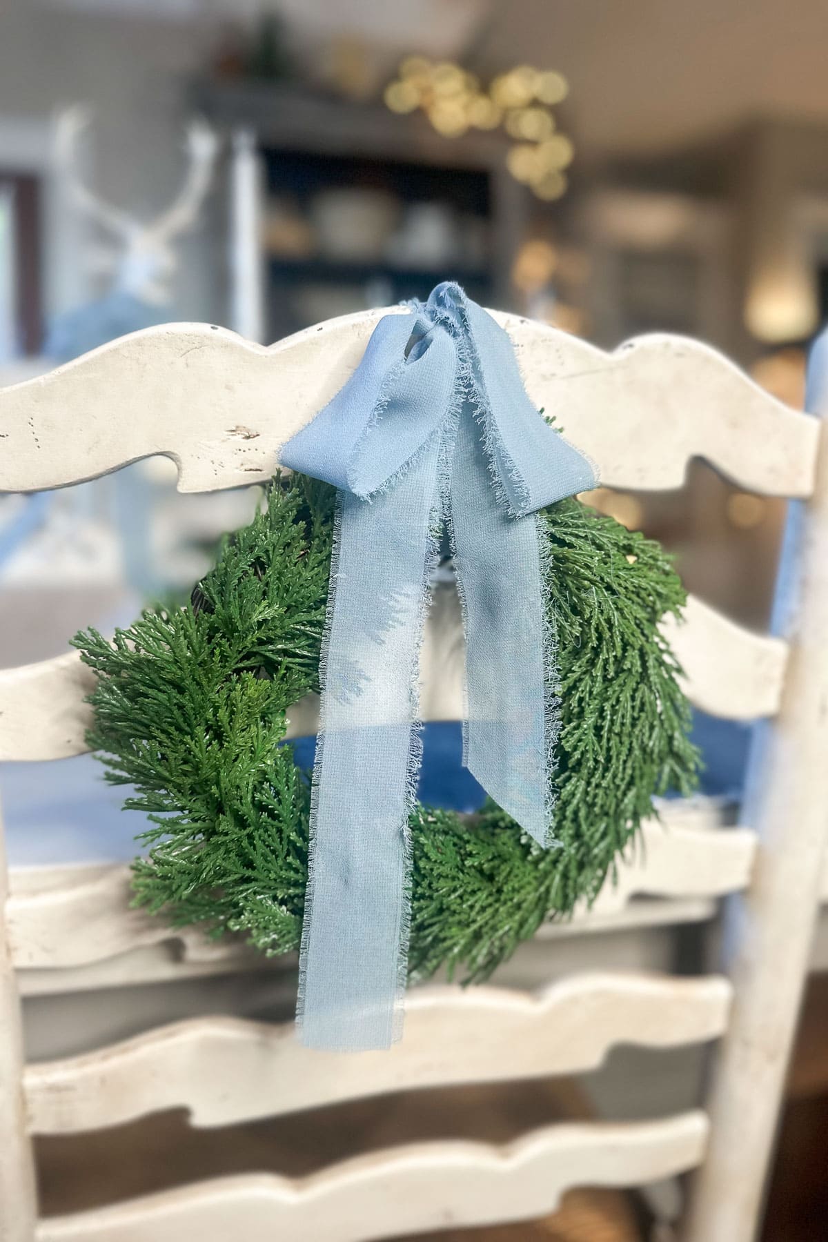 A small green wreath with a sheer blue ribbon hangs on the back of a white wooden chair. The background features a blurred interior with soft lighting, creating a cozy atmosphere.