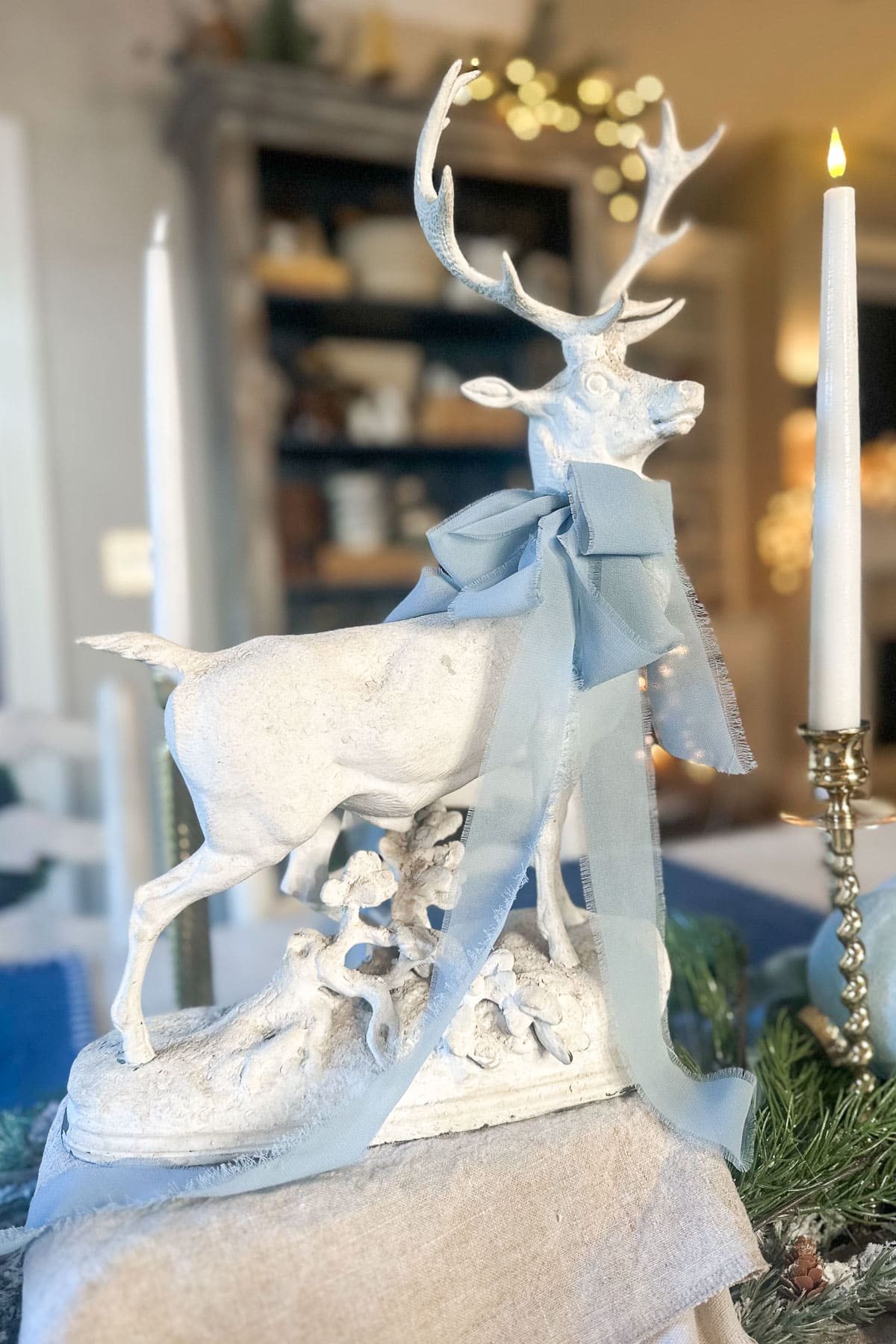 A decorative white deer statue with large antlers and a blue ribbon is placed on a table. Surrounding it are candles and greenery. The background features blurred kitchen shelves with subtle warm lighting.