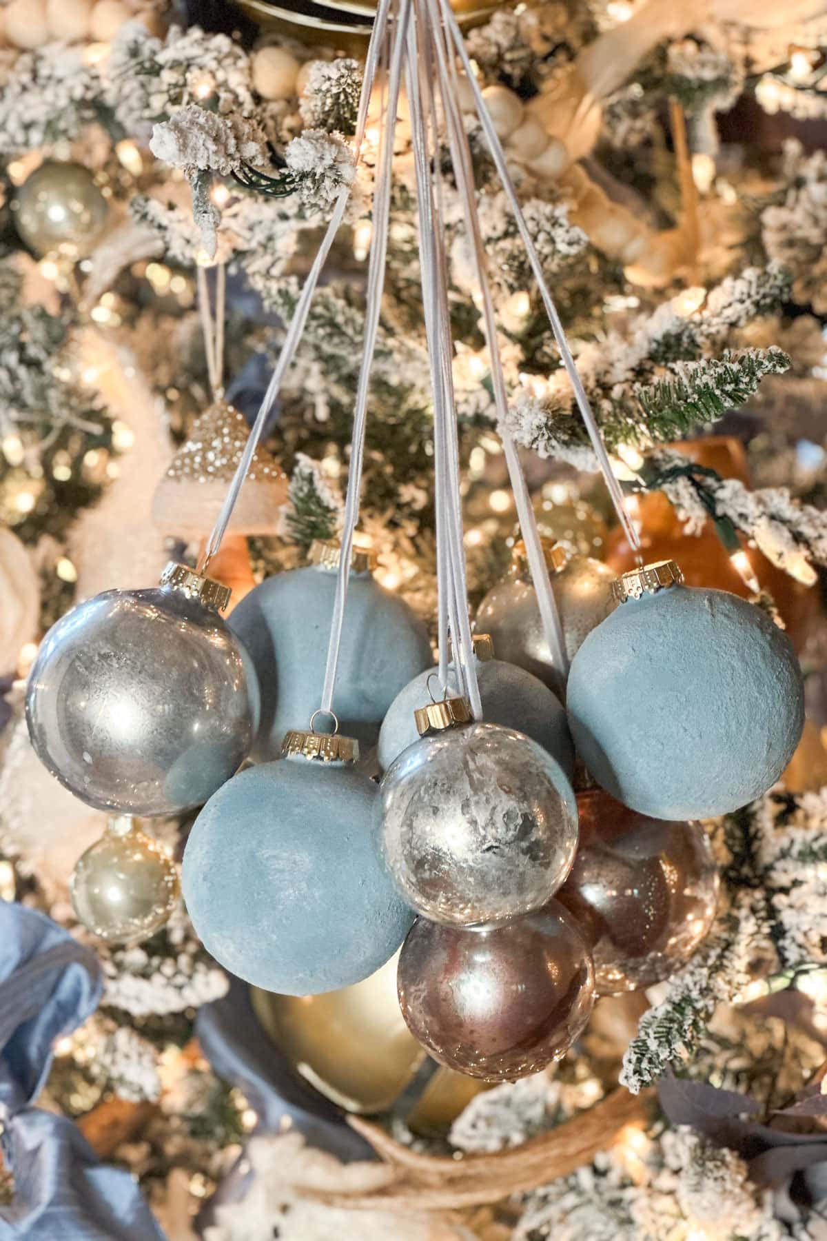 A cluster of blue and silver Christmas baubles hanging from a snowy, decorated tree branch. The tree is adorned with twinkling lights and other festive ornaments, creating a warm holiday atmosphere.