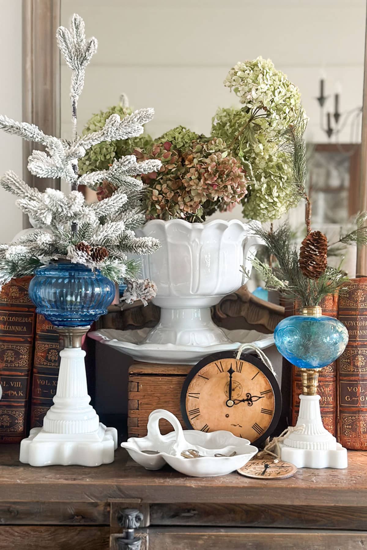 A decorative arrangement features vintage books, a clock, a white vase with dried hydrangeas, a small frosted faux tree, and two blue glass candle holders. All items are set on a wooden surface.