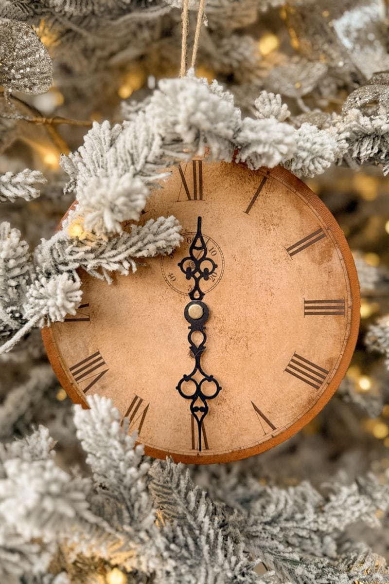 A vintage clock ornament with Roman numerals is nestled in a frosted, snow-covered Christmas tree. The clock hands point to 10:10. Twinkling lights add a warm glow to the scene.