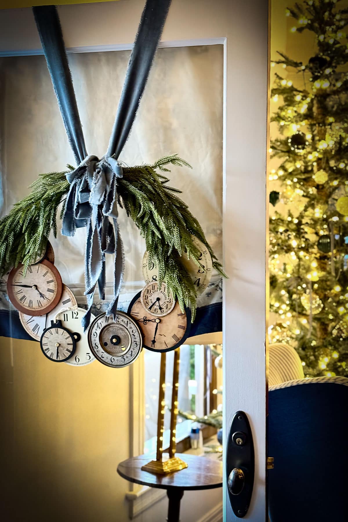 A door with a wreath made of various clock faces and green foliage, reminiscent of classic Christmas decor, hangs slightly open. Through the doorway, a warmly lit room is visible with an illuminated Christmas tree and a small table adorned with a glowing candle.