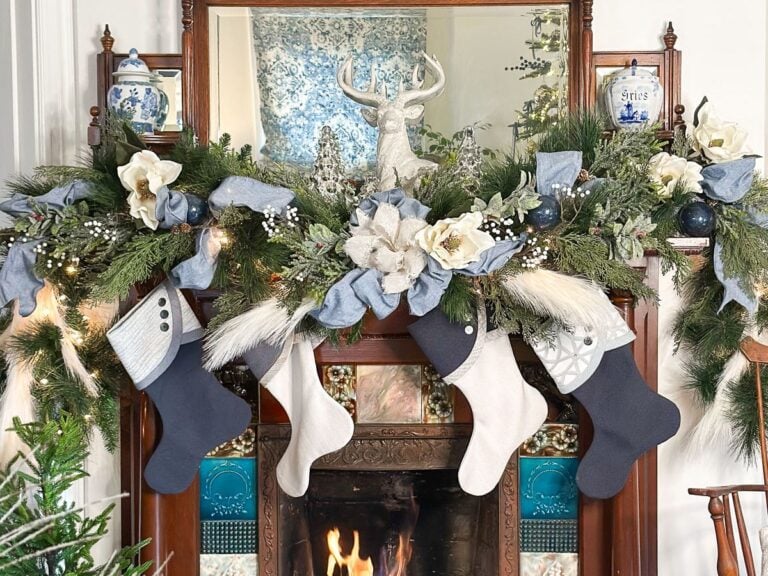 Festive mantle decorated with greenery, white flowers, and a reindeer figurine. Blue and white stockings hang below, above a lit fireplace. Blue-patterned ceramics accent the display, creating a cozy winter ambiance.