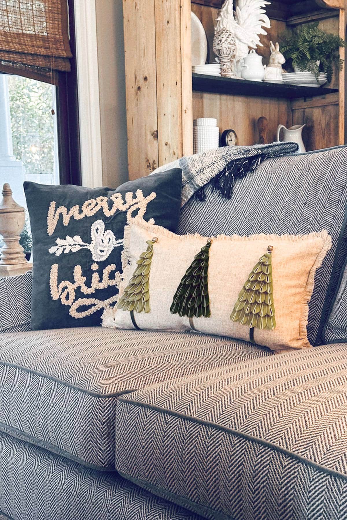 A cozy living room features a gray herringbone-patterned couch with two decorative pillows. One pillow reads "merry & bright" and the other has green velvet ribbon tree designs. There's a wooden bookcase and a window with blinds in the background.