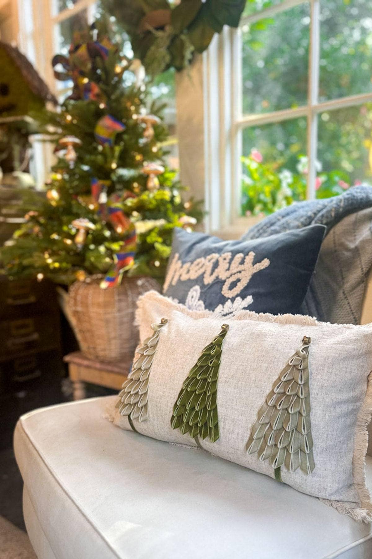 A cozy living room scene with a small decorated Christmas tree by a window. A white couch features two decorative pillows, one with green tree designs and another with the word u0022Merryu0022 on it. Sunlight streams through the foliage outside.