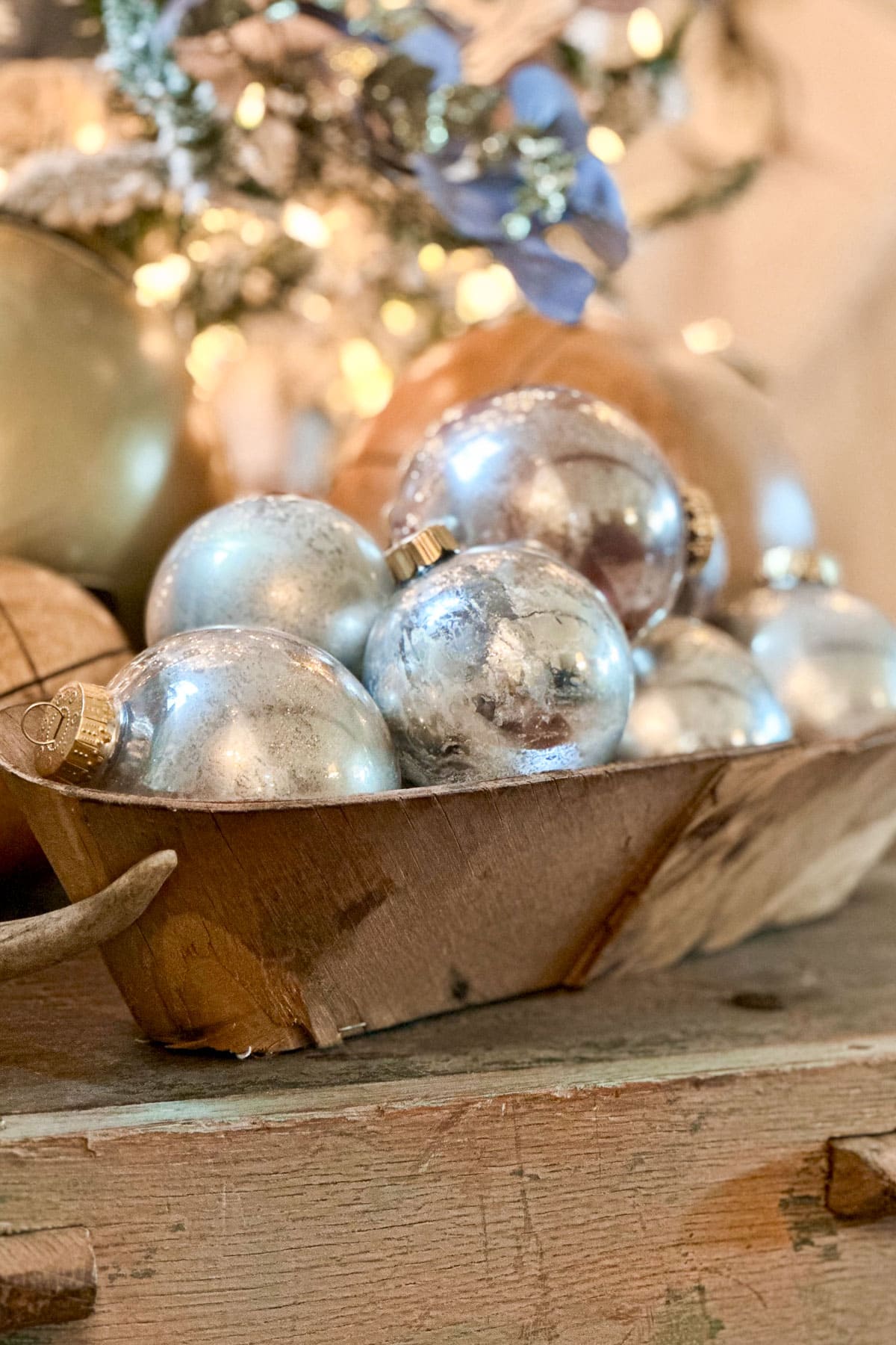 A wooden tray filled with DIY mercury glass ornaments is displayed on a rustic wooden surface. In the blurred background, a decorated Christmas tree with lights and a blue ribbon adds a festive glow.