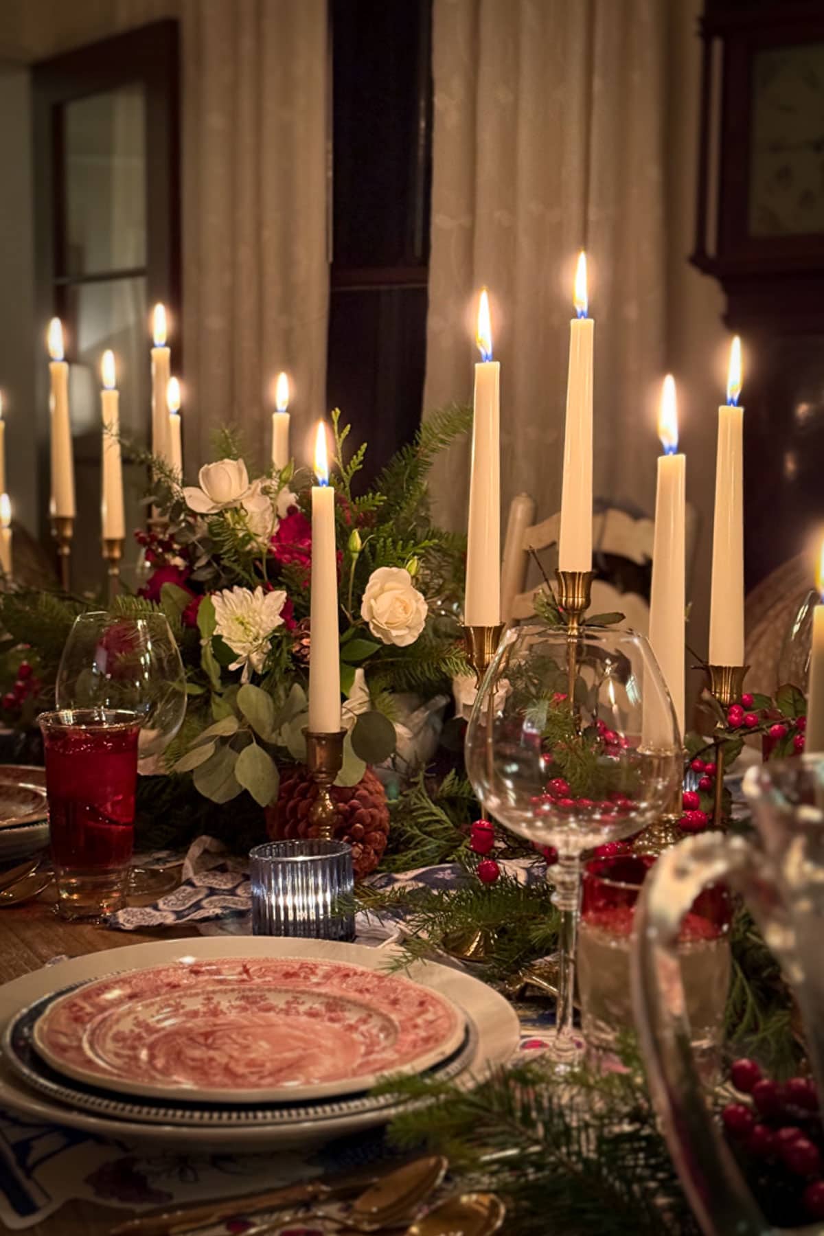 A warmly lit dining table exudes Classic Christmas Decor, ready for a festive gathering. Tall candles and floral arrangements with white roses, red berries, and greenery adorn the scene. Plates are neatly arranged, with wine glasses reflecting the candlelight's glow.