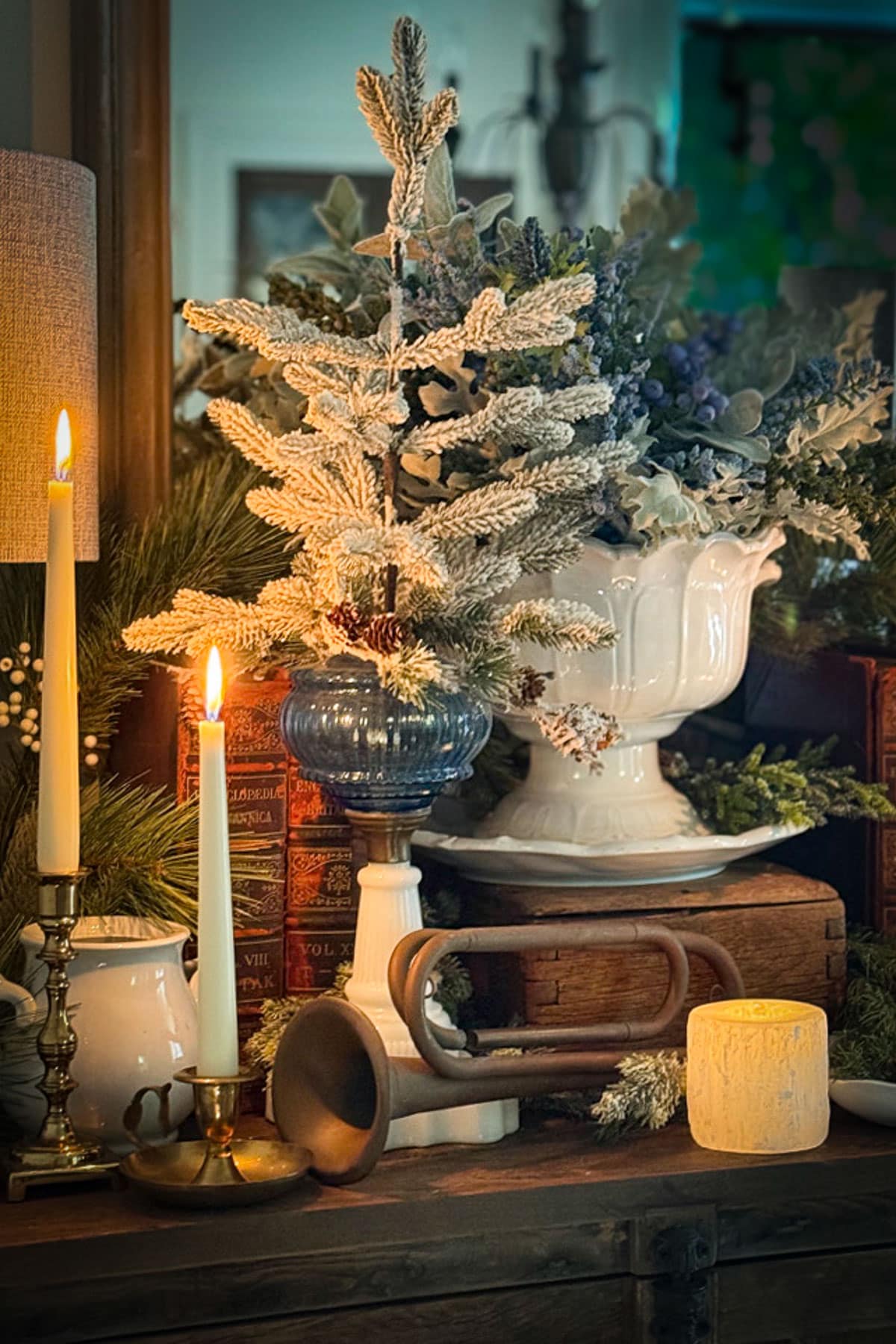 A cozy, vintage-style display features lit candles, a frosted mini Christmas tree in a blue vase, a white pitcher, antique books, and a decorative horn on a wooden table, creating a warm and festive atmosphere.