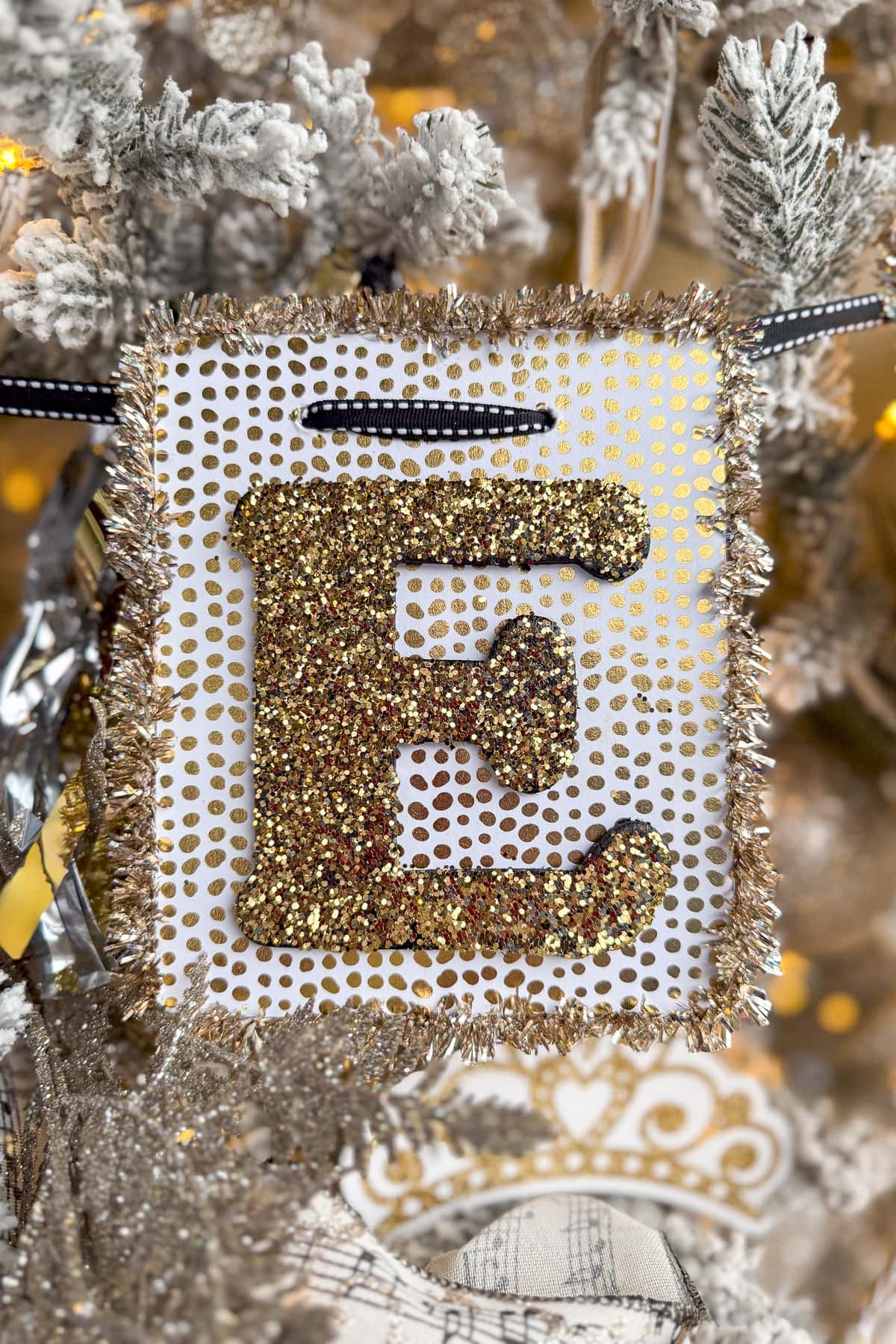 A decorative ornament shaped like the letter "E" with gold glitter and a sparkling border hangs on a snow-dusted New Year's tree. Warm yellow lights illuminate the background, setting the festive scene aglow.