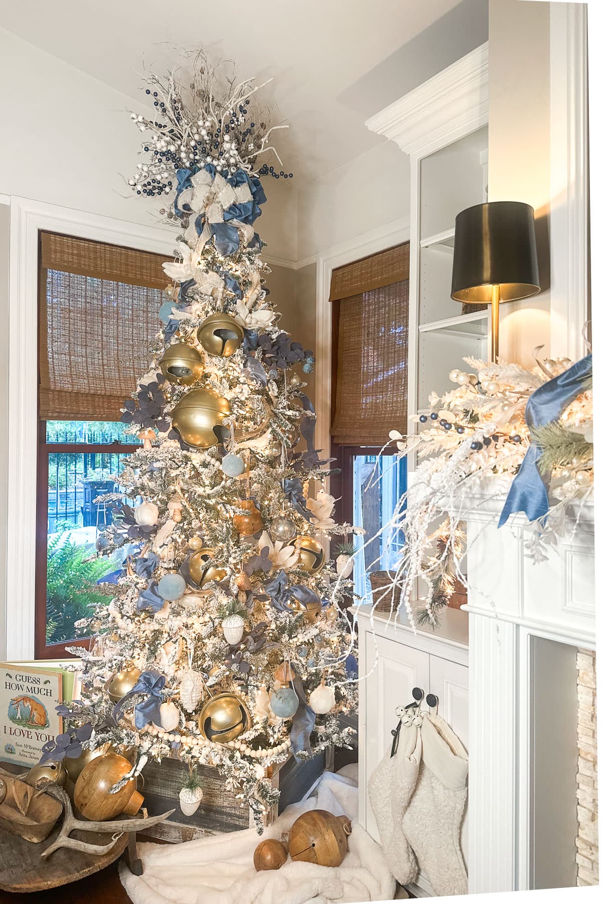 A tall, white Christmas tree adorned with blue and gold ornaments and ribbons stands next to a fireplace. Embracing the blue and white Christmas decor theme, stockings hang on the mantel, and gifts are placed around the tree on a white rug. The room features large windows with brown blinds.