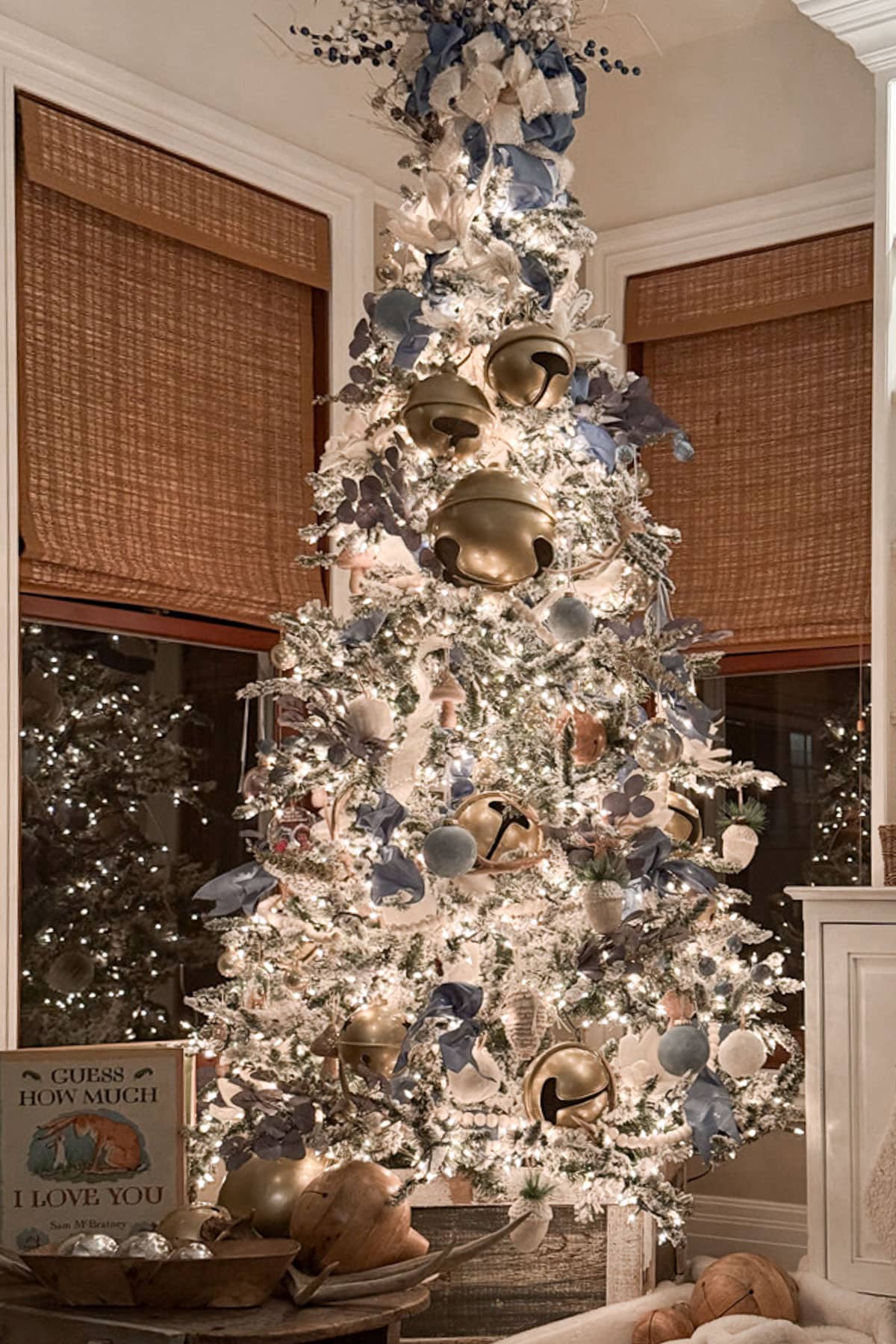 A beautifully decorated Christmas tree featuring blue and white Christmas decor, adorned with shimmering white lights, blue and gold ornaments, and large golden bells at the center. A sign nearby reads, "Guess How Much I Love You." The tree is set in a cozy room with wooden blinds.