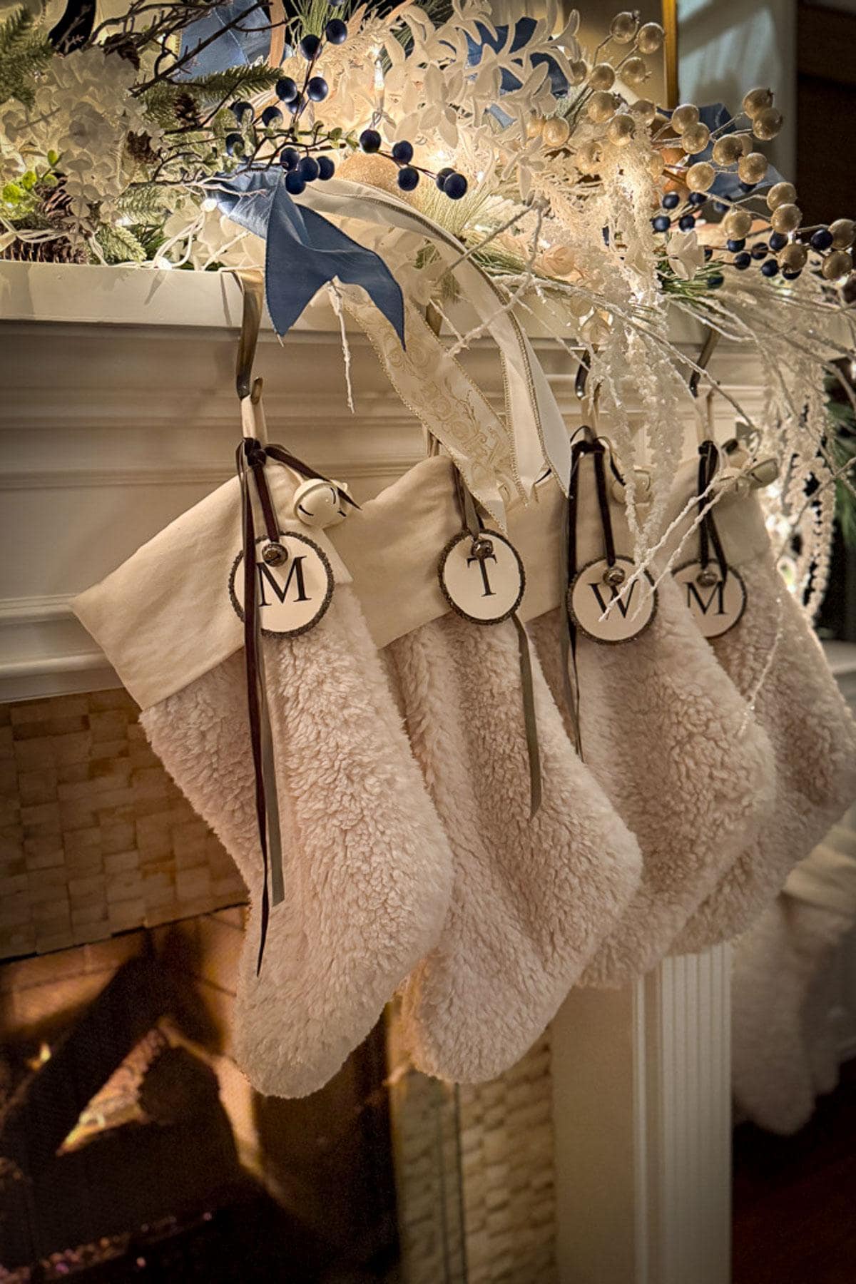 Four fluffy stockings hang from a decorated mantel, each with the initials "M," "T," "W," and "M" on wooden tags. The fireplace is lit, and the classic Christmas decor of white and blue festive ornaments adorns the mantel.