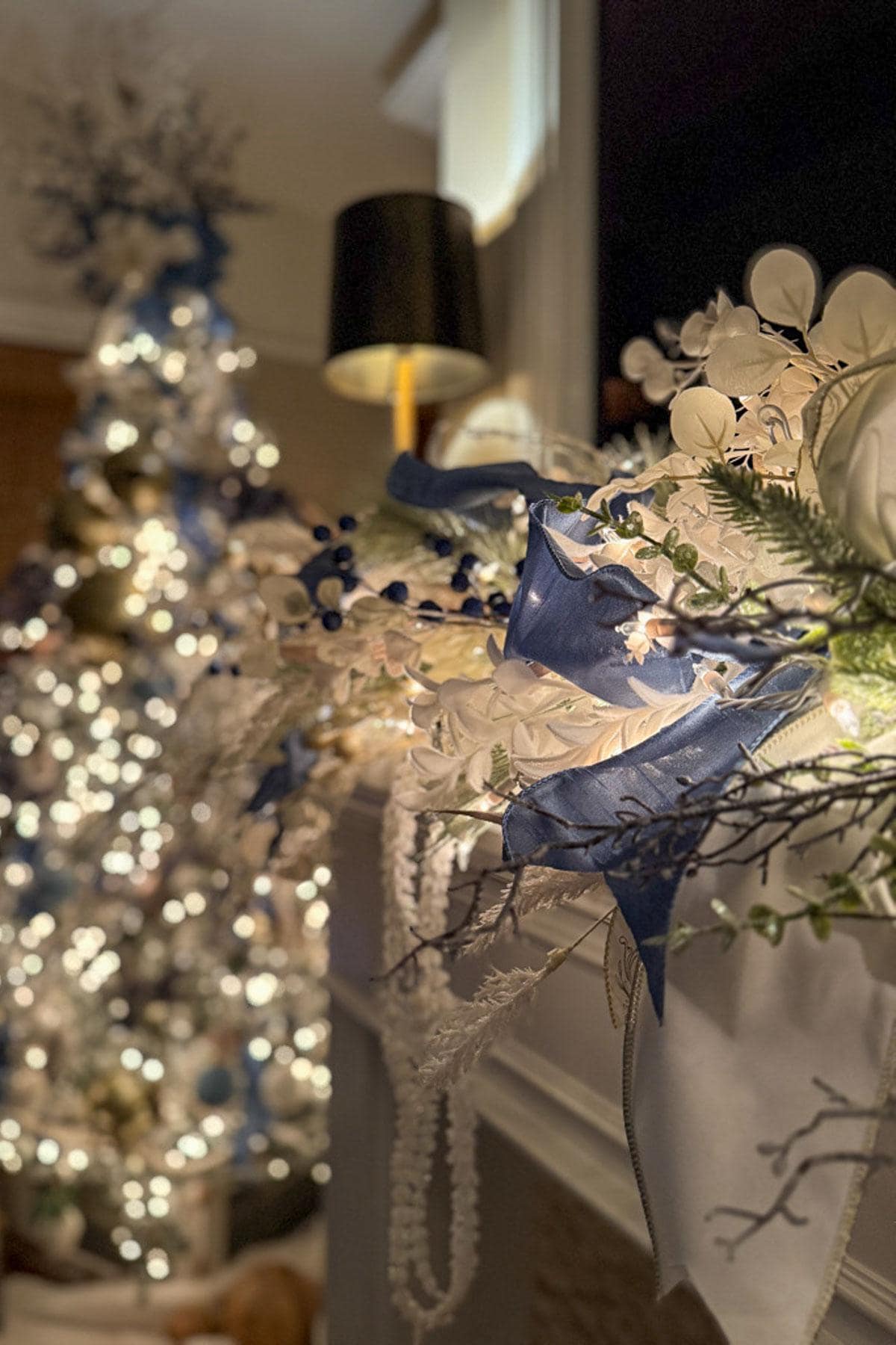 A cozy room epitomizing classic Christmas decor features a glowing tree in the background. In the foreground, a mantel is adorned with white and blue flowers, berries, and soft lights. A black lamp provides additional warm lighting.