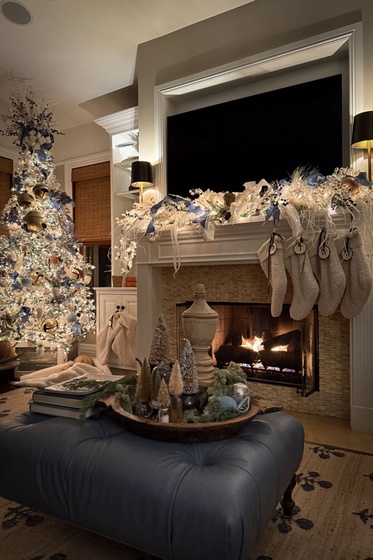 A cozy family room with classic Christmas decor features a beautifully decorated tree, stockings hanging on a lit fireplace, and a tray of holiday decor on a blue ottoman. The warm, festive atmosphere is enhanced by the glow of the fire and soft lighting.