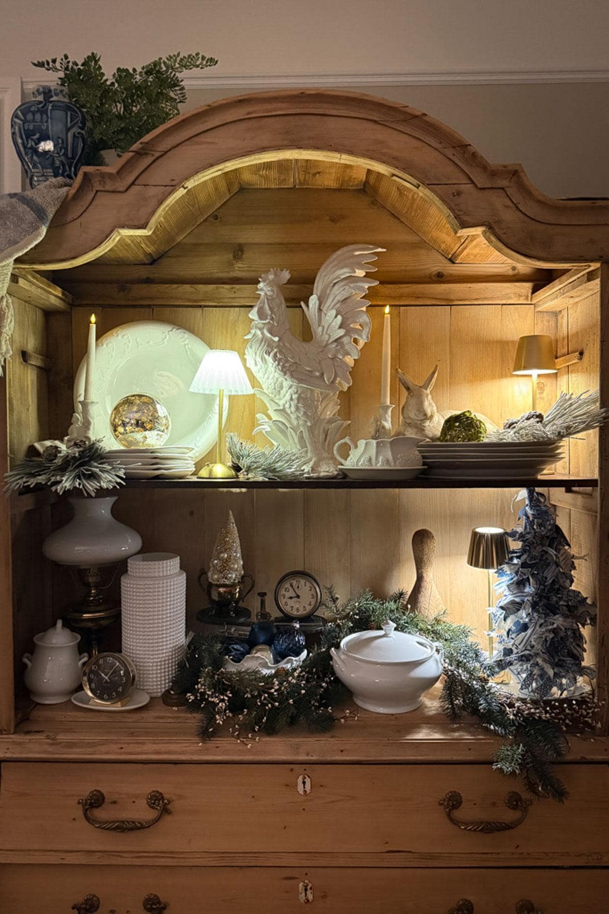 A wooden cabinet showcases classic Christmas decor with a large white rooster sculpture, candles, small lamps, stacked plates, a clock, and greenery for a festive look. The scene is warmly lit, creating a cozy atmosphere perfect for the holiday season.