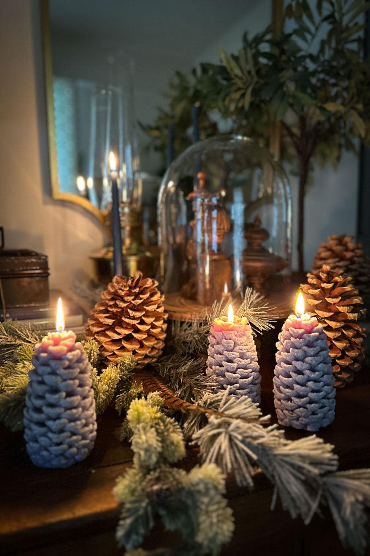 A cozy scene features three lit candles shaped like pine cones on a wooden table surrounded by pine branches and natural pine cones. Classic Christmas decor comes alive in the background, where a mirror reflects soft lighting and decorative items rest under a glass dome.