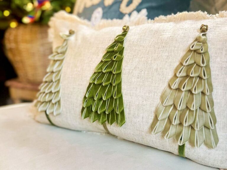 A decorative pillow with a cream fabric background features three green ribbon trees. Each tree is made from layered ribbon strips, creating a textured, leafy appearance. The pillow adds a festive touch to the setting.