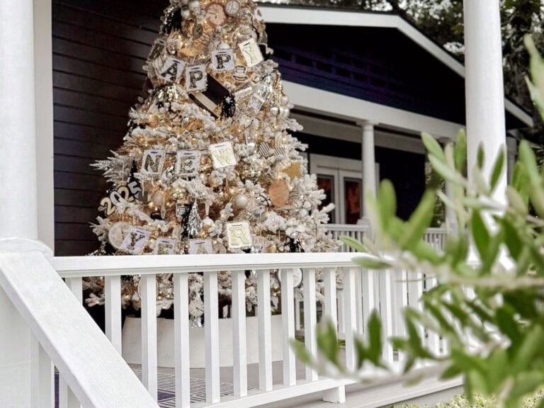 A festive white Christmas tree on a porch, adorned with silver and gold New Year's decorations, and large letters spelling "Happy New Year." The background showcases a dark blue house, while green leaves peek through in the foreground.