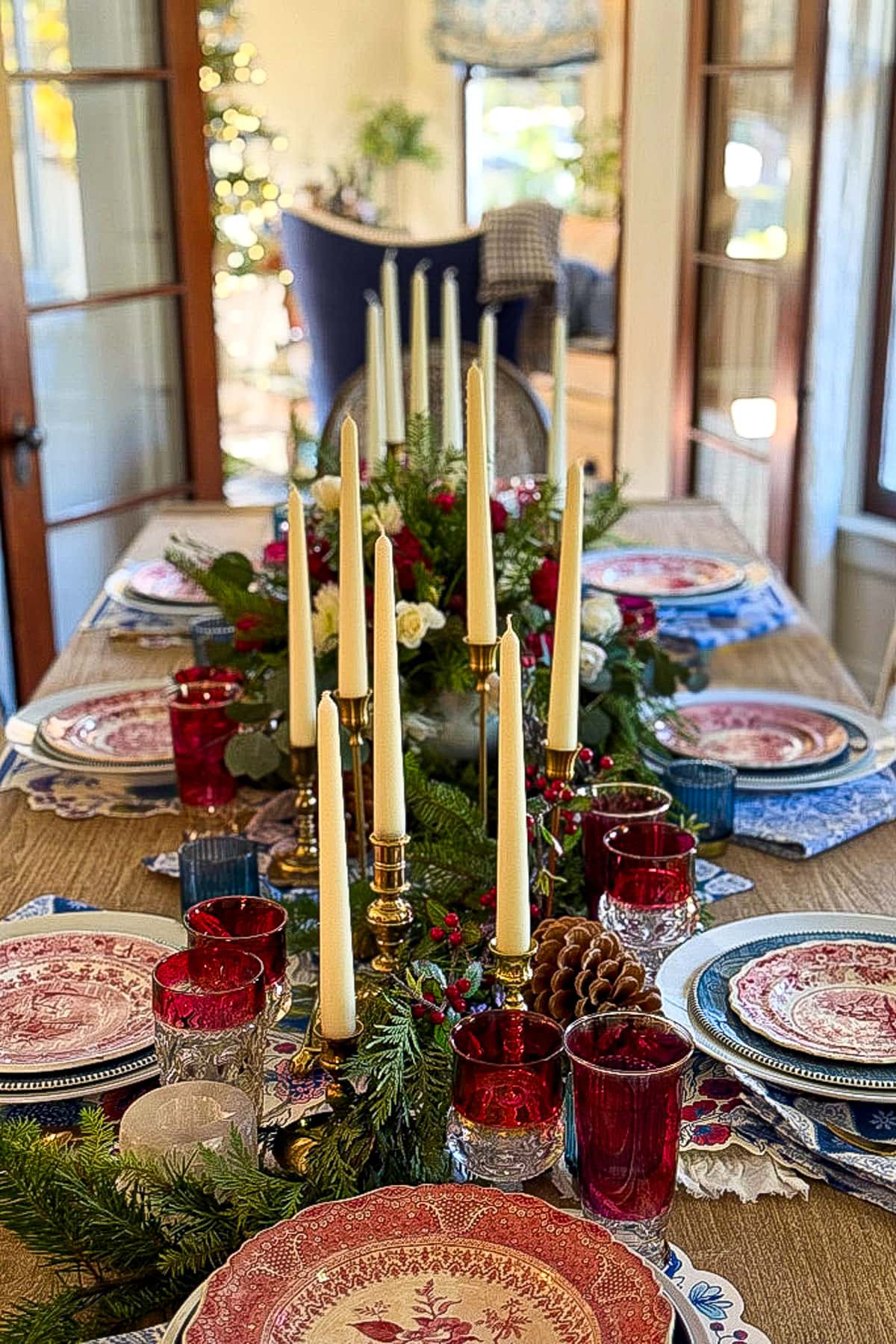 A festive dining table set for a holiday meal embodies classic Christmas decor with red and blue plates, red glassware, tall ivory candles, and a lush centerpiece of greenery and flowers. In the background, a decorated Christmas tree is visible near a window.