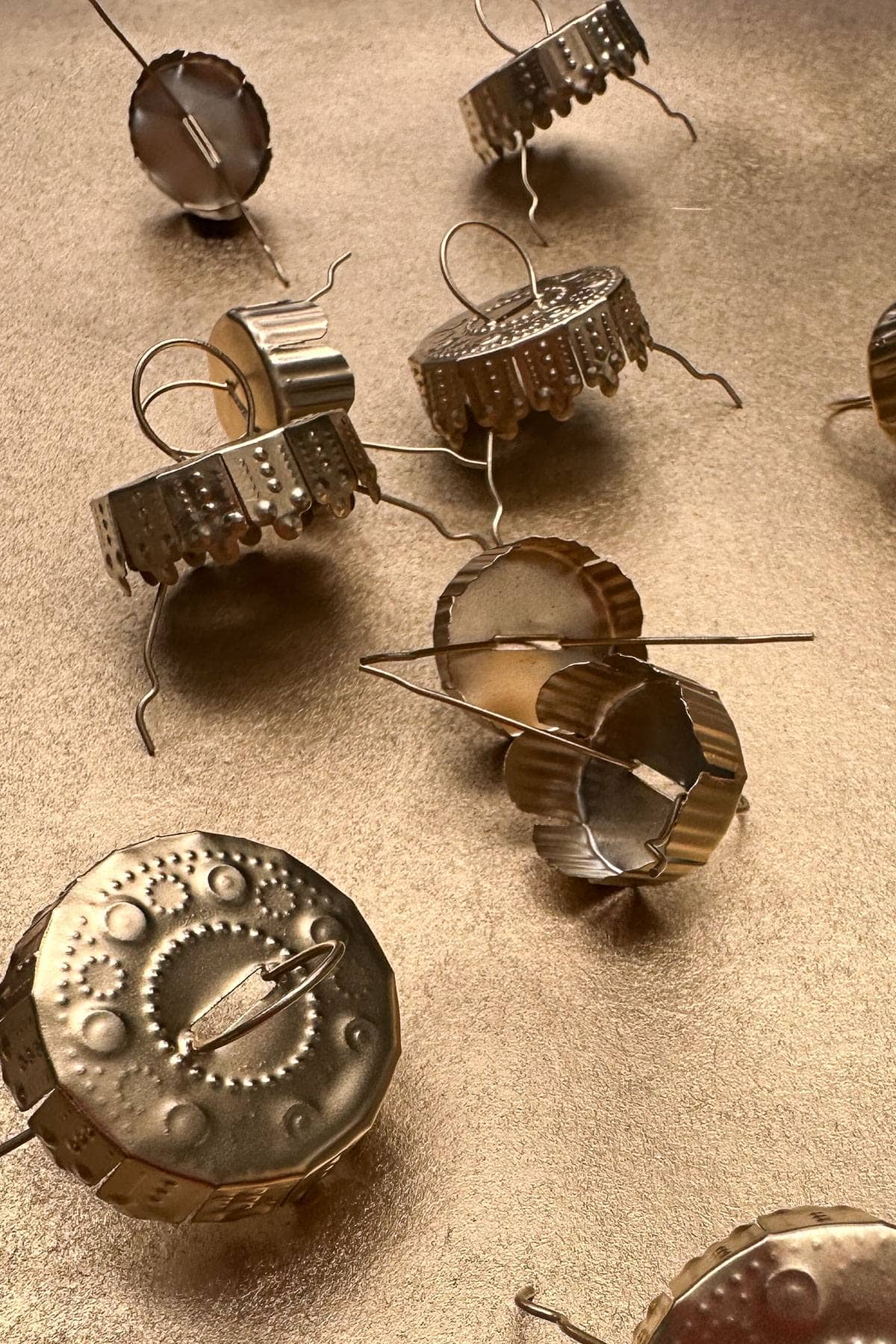 A collection of metallic, round pins with intricate designs scattered on a textured, bronze surface. The pins have various patterns and protruding rods, creating a decorative, artistic arrangement.