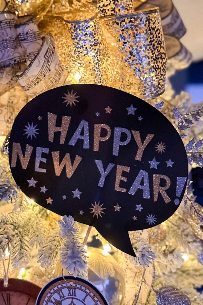 A festive Christmas tree adorned with gold ribbon and decorations. A speech bubble sign on the tree reads "Happy New Year!" in glittery letters. Sparkling stars and a clock ornament add to the celebratory ambiance.