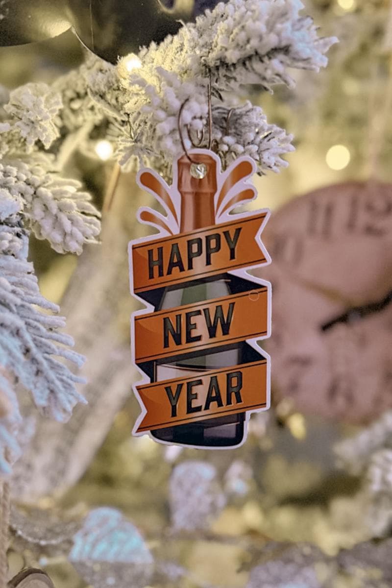 A festive holiday ornament shaped like a bottle, with the text "Happy New Year" in bold orange and black displayed on it. The ornament hangs on a snow-dusted Christmas tree branch, with warm lights and a clock blurred in the background.