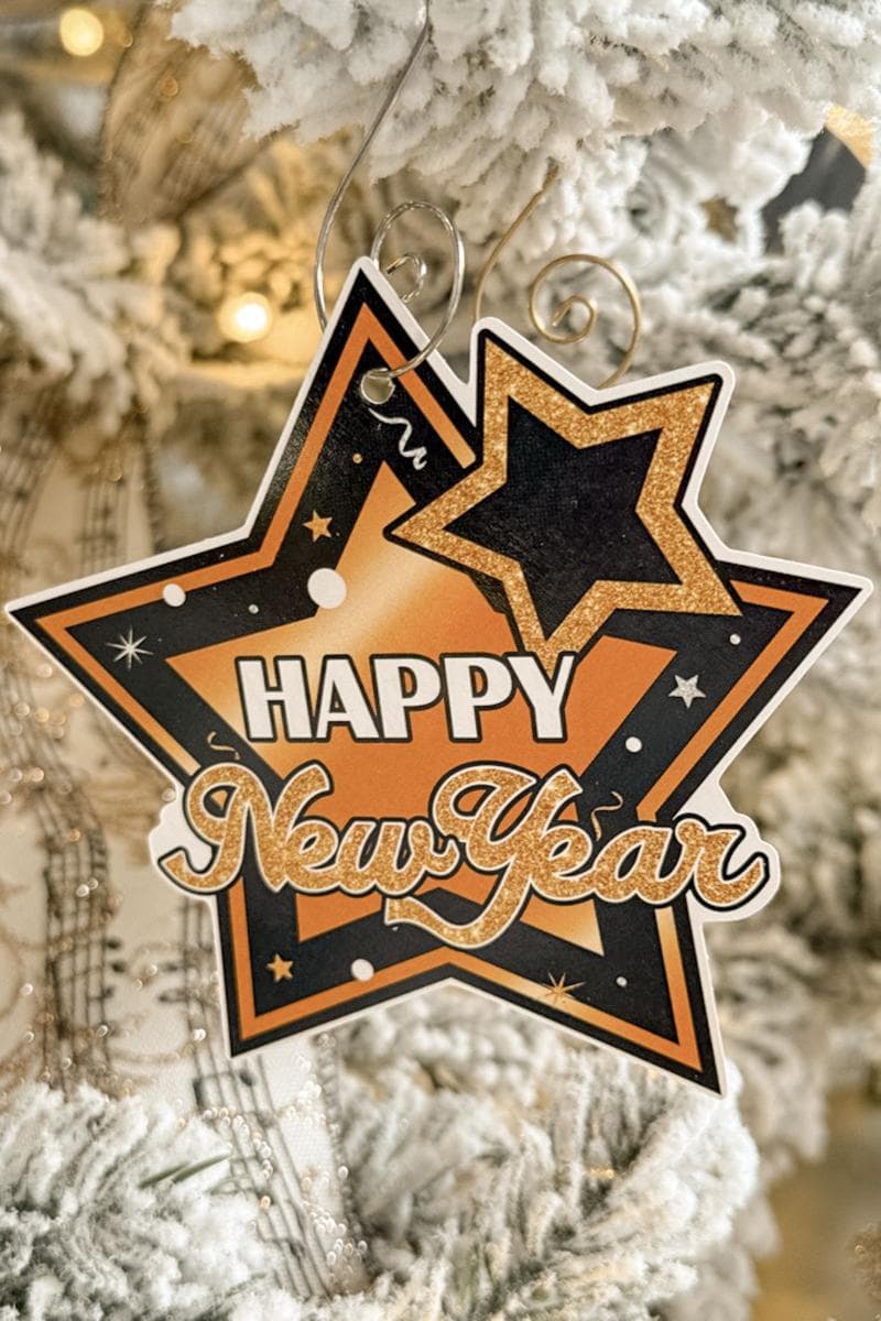 A festive "Happy New Year" ornament hangs on a snowy Christmas tree. It features a black and gold star design with celebratory confetti motifs.