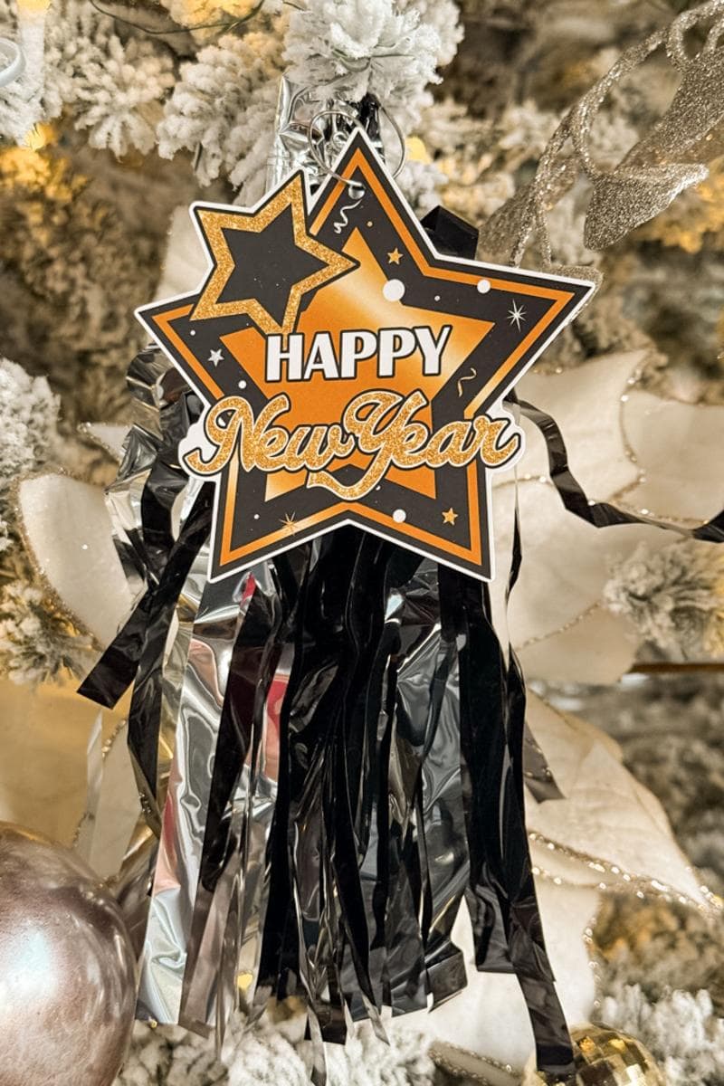 A sparkling New Year decoration hangs on a snowy Christmas tree branch. The ornament is shaped like a star and reads "Happy New Year" in metallic gold and black. Shiny black and silver streamers dangle below.