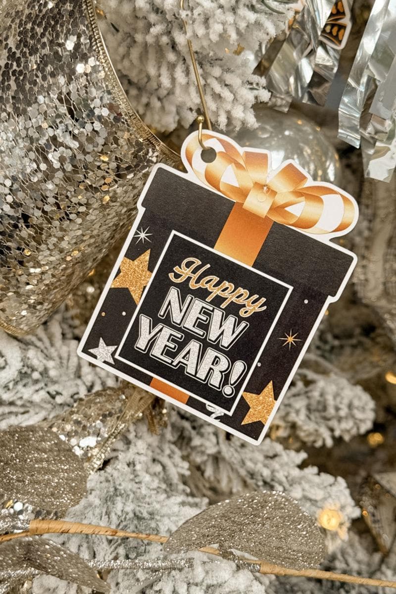 A festive close-up of a snowy Christmas tree adorned with gold and silver ribbons. A black gift tag with gold stars displays the message "Happy New Year!" in bold letters.