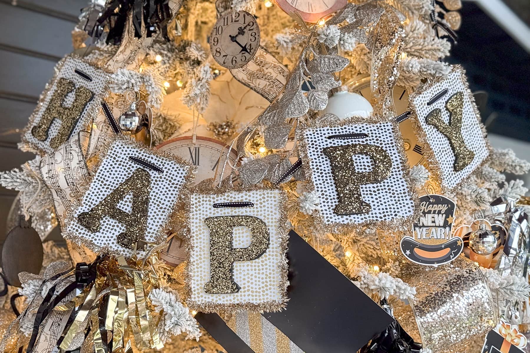 A festive New Years tree adorned with sparkling lights and decorations, featuring large, glittery letters that create a banner spelling "HAPPY." A small "Happy New Year" ornament is also visible. The tree has a mix of gold, silver, and white embellishments.