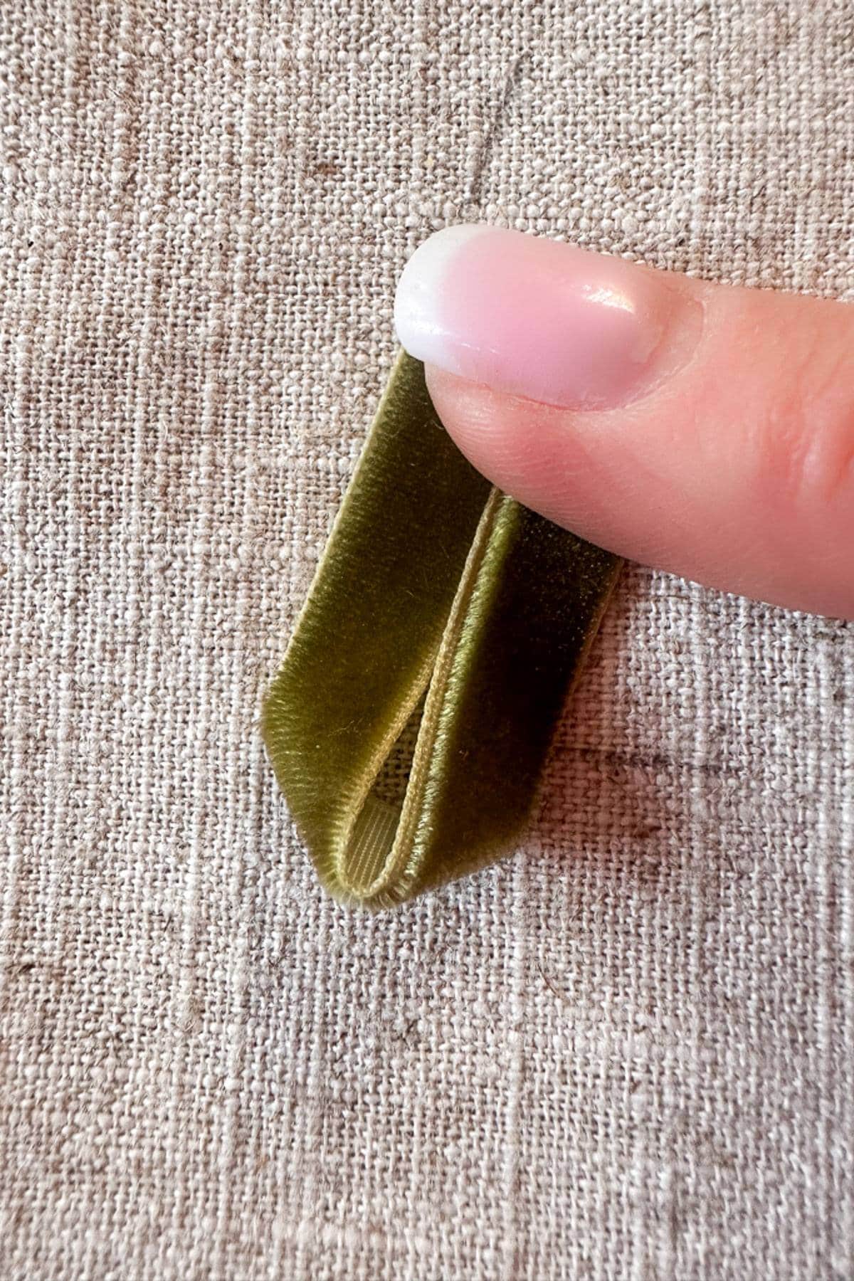 A hand with pink nails pinching a small loop of green velvet ribbon against a textured beige fabric background.