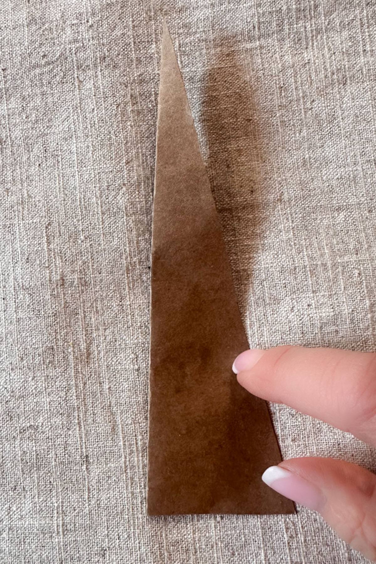 A tree template of brown paper is placed on a pillowcase surface. A hand with neatly manicured nails is lightly touching the edge of the paper. a Shadows are visible on the fabric.