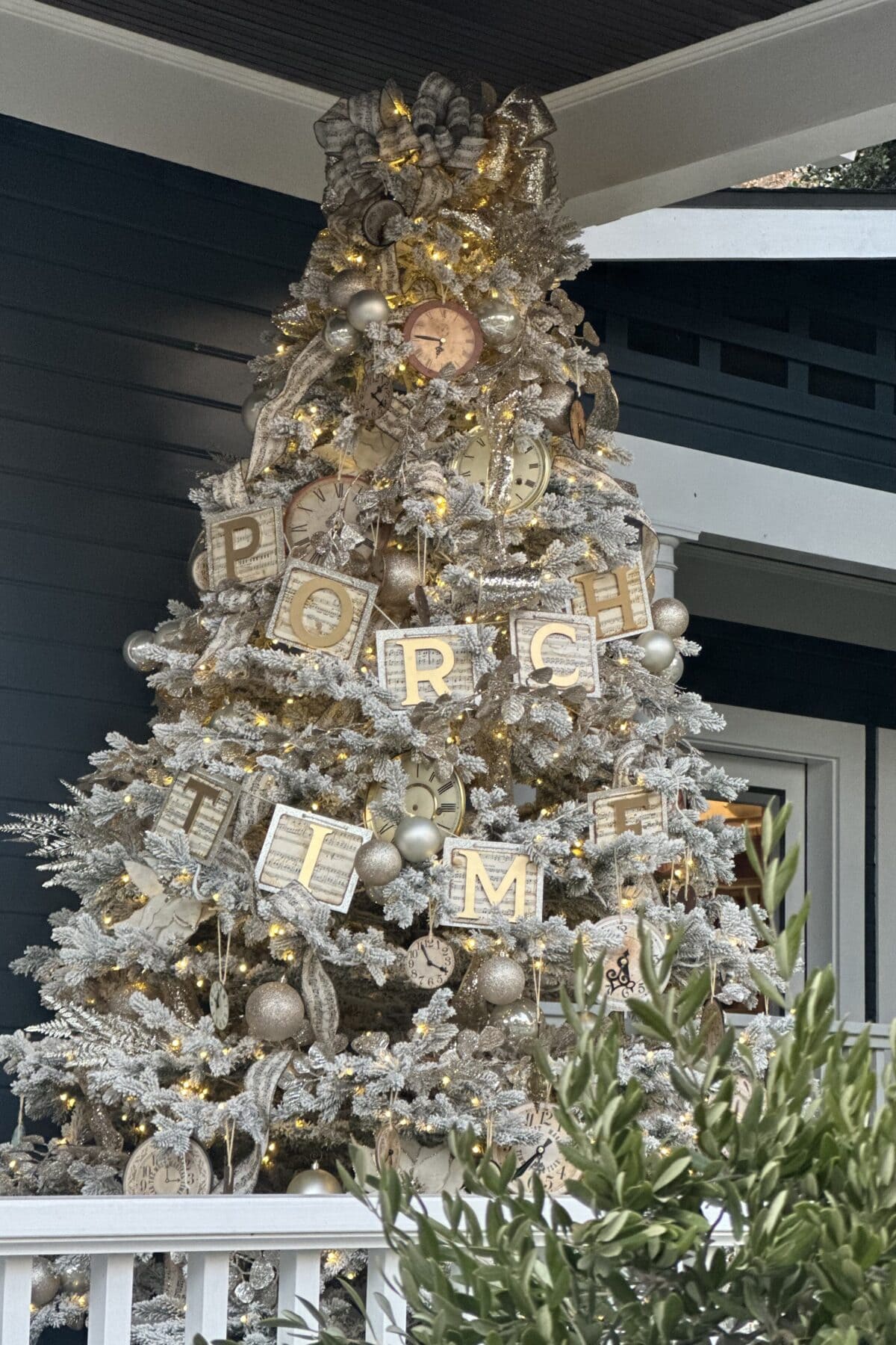 A Christmas  tree on a porch, adorned with clocks, ornaments, and large letters spelling "PORCH TIME." The tree is dusted with artificial snow and surrounded by greenery. Celebrate the season with these unique decorations that blend Christmas charm with New Year cheer.