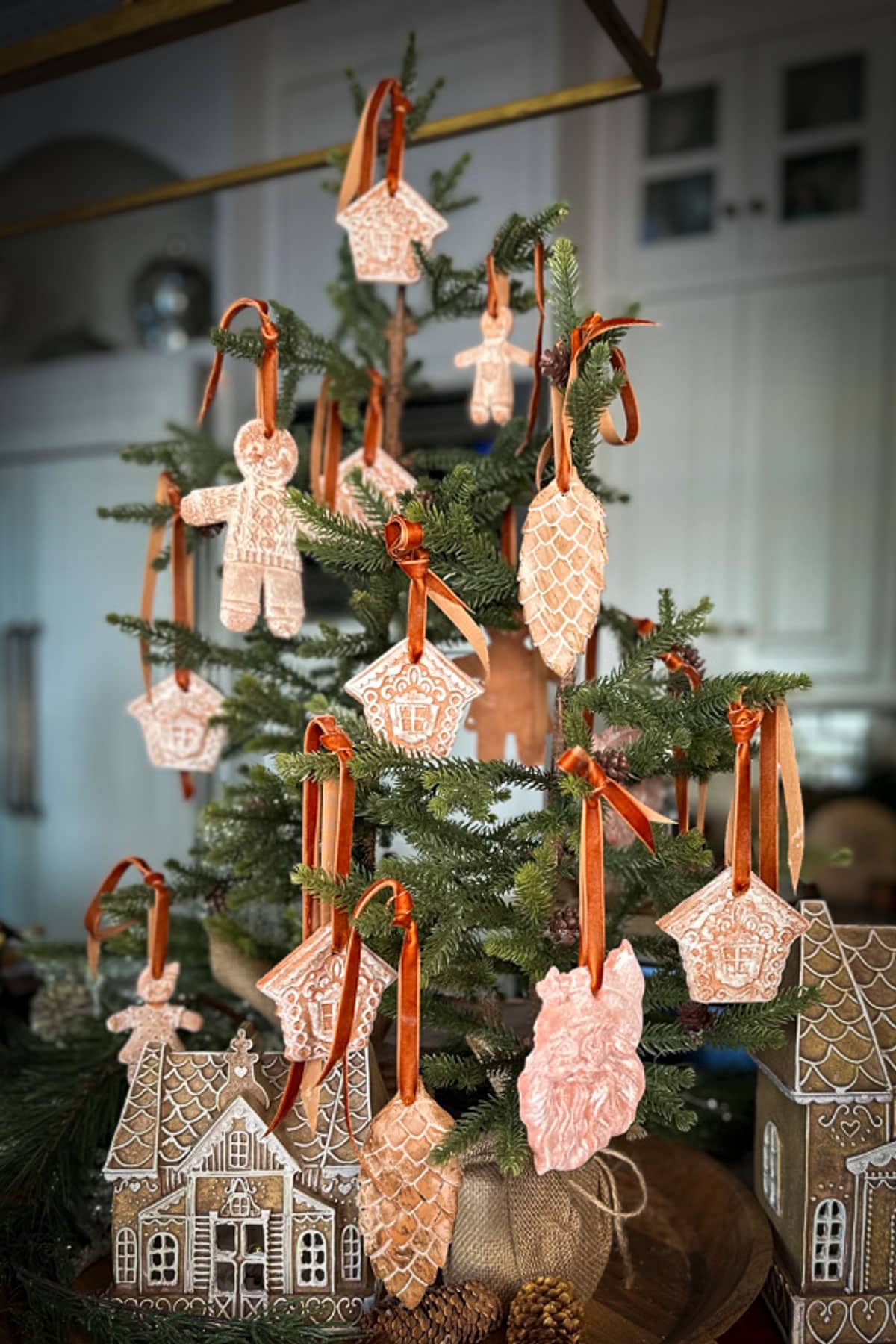 A small Christmas tree, embodying classic Christmas decor, is adorned with gingerbread-themed ornaments, including houses, pinecones, and figures hanging from orange ribbons. The tree is set on a table alongside decorative ceramic houses in a gingerbread style.