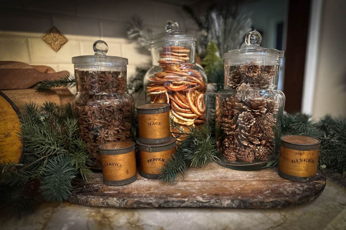 A wooden tray displays glass jars filled with star anise, dried citrus slices, and pinecones. Small vintage spice tins labeled "Cloves," "Pepper," and "Ginger" are arranged in front. Evergreen branches add a festive touch to the cozy kitchen scene.