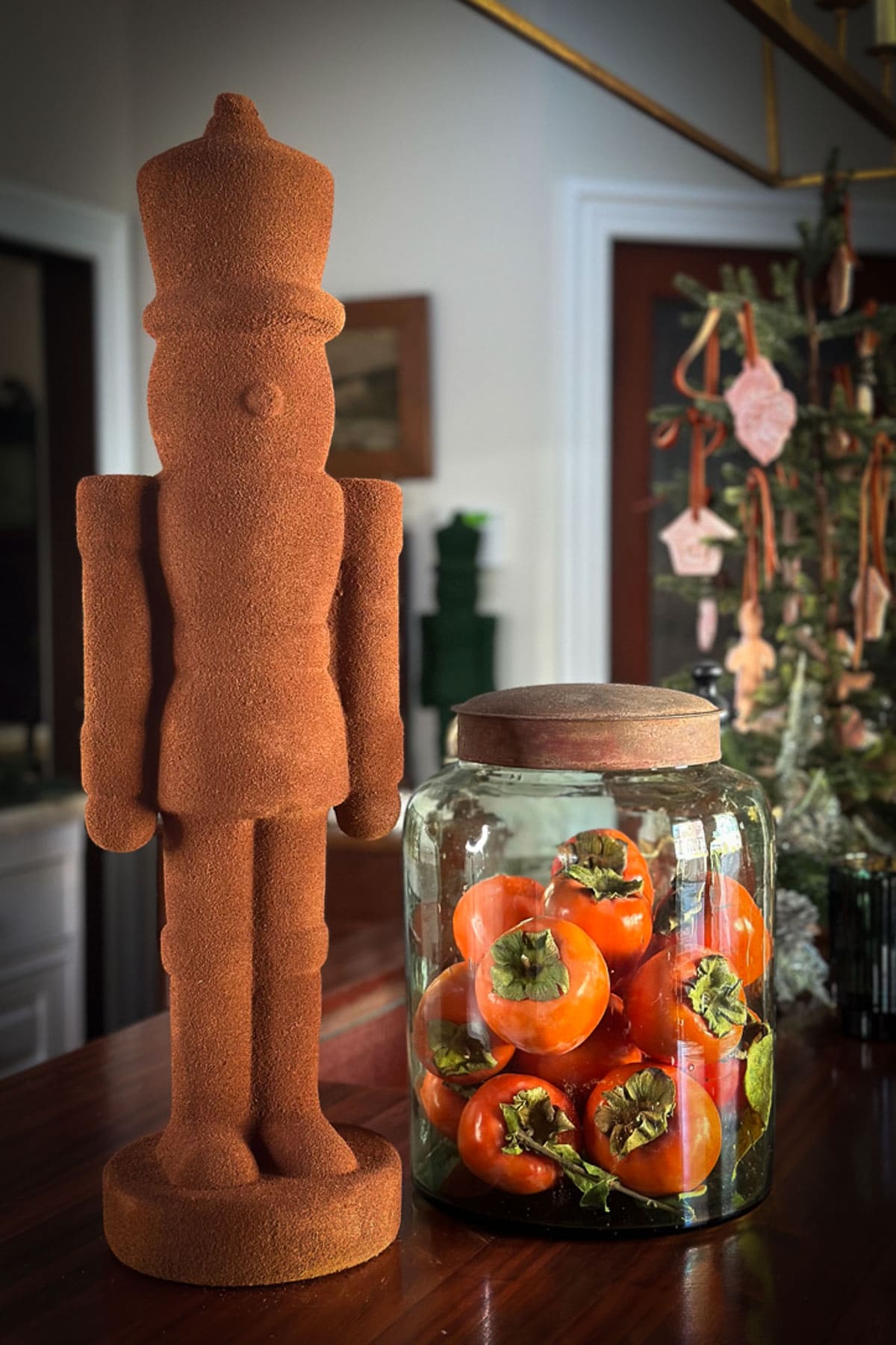 A rustic nutcracker figurine, a staple of classic Christmas decor, stands beside a glass jar filled with ripe persimmons on a wooden table. In the background, a small decorated Christmas tree enhances the festive spirit near a doorway.