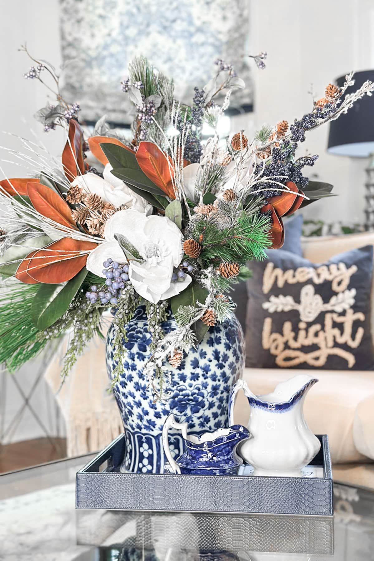 A decorative blue and white vase with an arrangement of white flowers, pinecones, and greenery sits on a tray. There's a matching ceramic pitcher and creamer nearby. In the background, a pillow with "merry & bright" is on a couch.