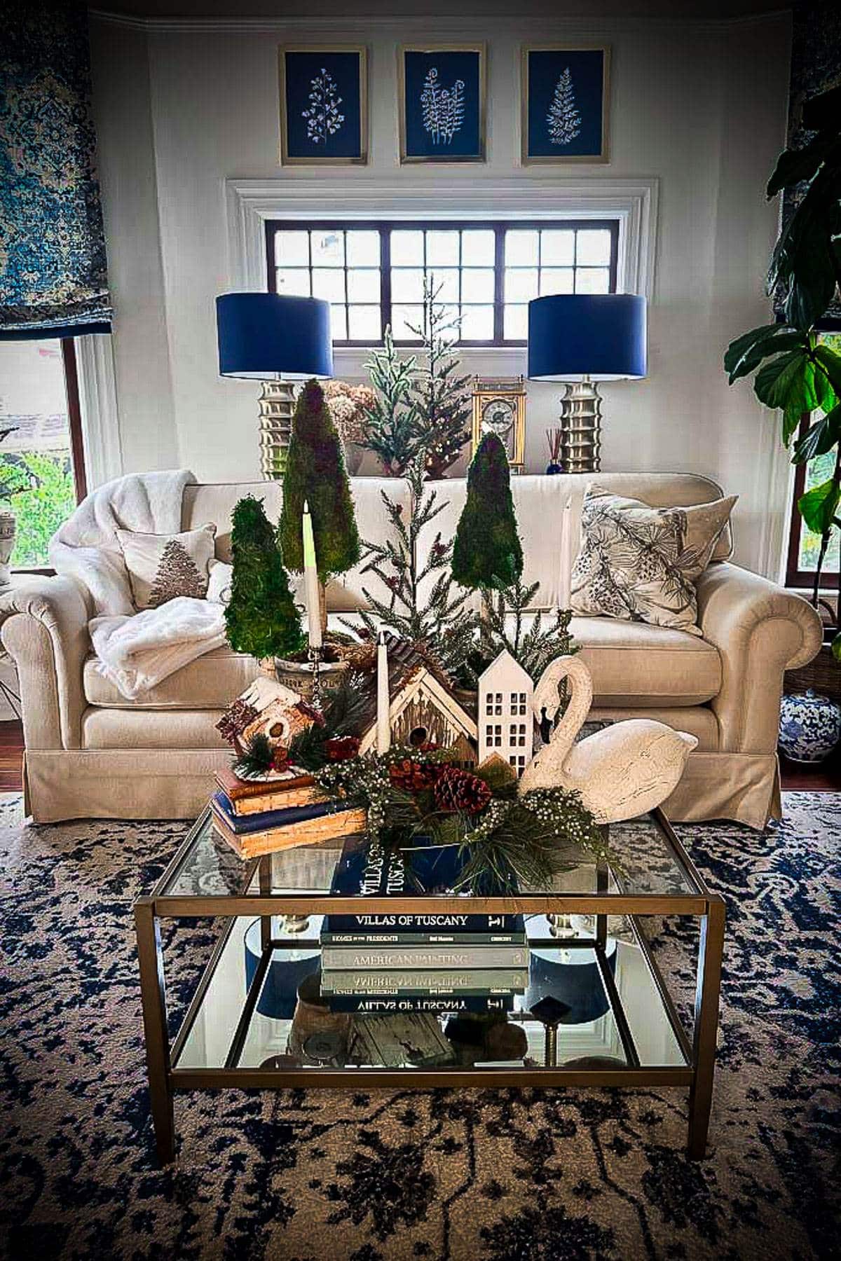 A cozy living room features a cream sofa with patterned pillows, a coffee table with books and decorative items like candles and miniature trees. Two blue lamps flank a window above the sofa, and framed botanical prints adorn the wall.