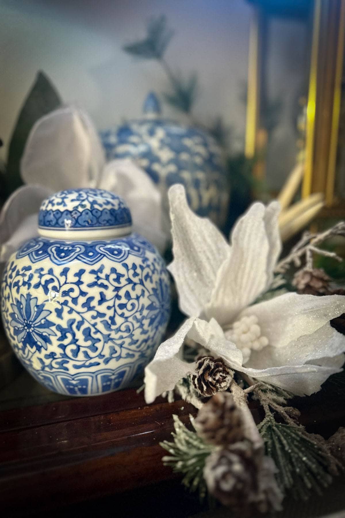 A decorative scene featuring a blue and white intricately patterned porcelain jar, surrounded by white flowers, pine cones, and green foliage with a soft focus background.