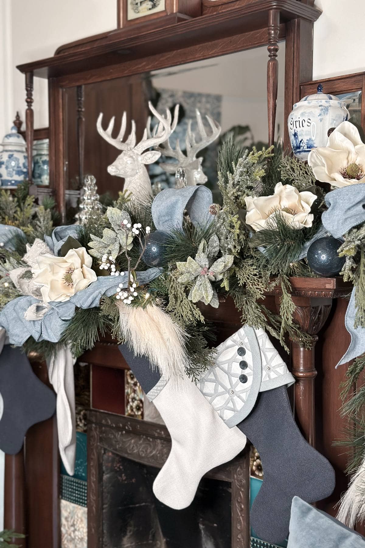 A festive mantel decorated for the holidays features a mix of blue and white stockings, greenery, flowers, and ornaments. A white reindeer figurine is centered, flanked by blue and white vases and large magnolia flowers.