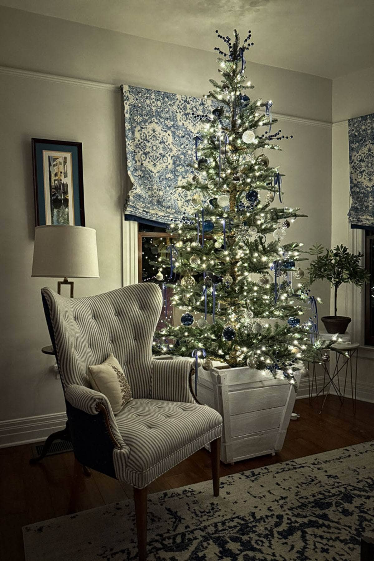 A cozy living room features a tall, decorated Christmas tree with white and blue ornaments and lights. An elegant, striped armchair with a small cushion is placed nearby. The room has patterned blue and white curtains and a framed picture on the wall.