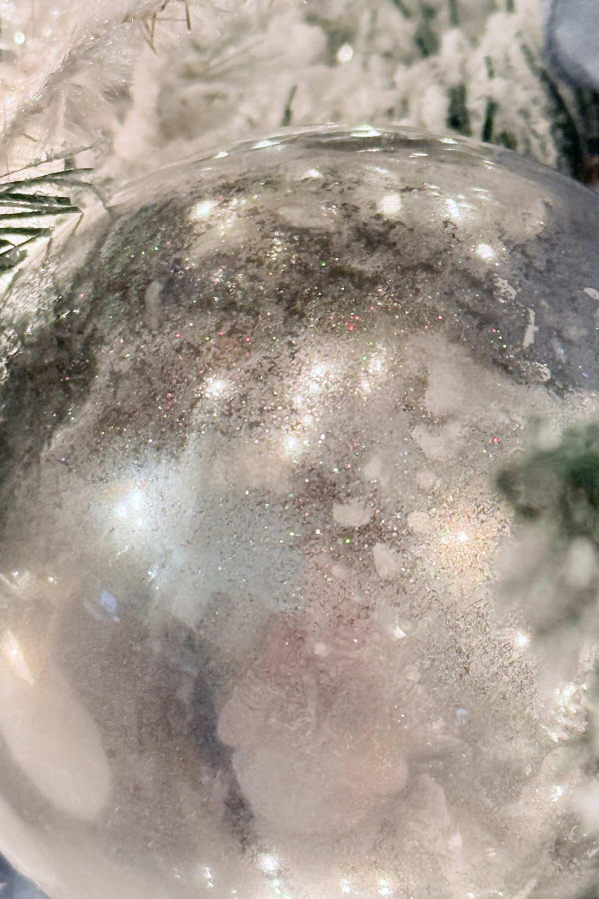 diy mercury glass ornaments-A close-up of a DIY Mercury Glass Ornament with a shimmering, reflective surface. It hangs on a snow-dusted Christmas tree, capturing glints of festive lights and green foliage in the background.