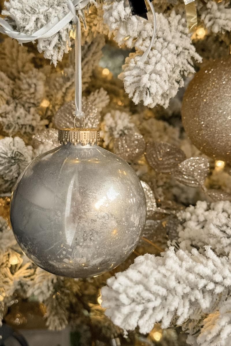 A close-up of a Christmas tree adorned with snow-covered branches and a large silver ornament hanging from a white ribbon. The background features warm white lights and sparkling gold decorations.