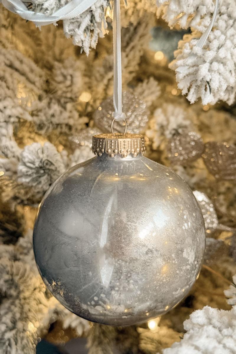 A silver Christmas ornament hangs from a frosted evergreen tree branch. The ornament reflects light, surrounded by soft, white snow-like decorations and warm glowing lights, creating a festive and cozy atmosphere.