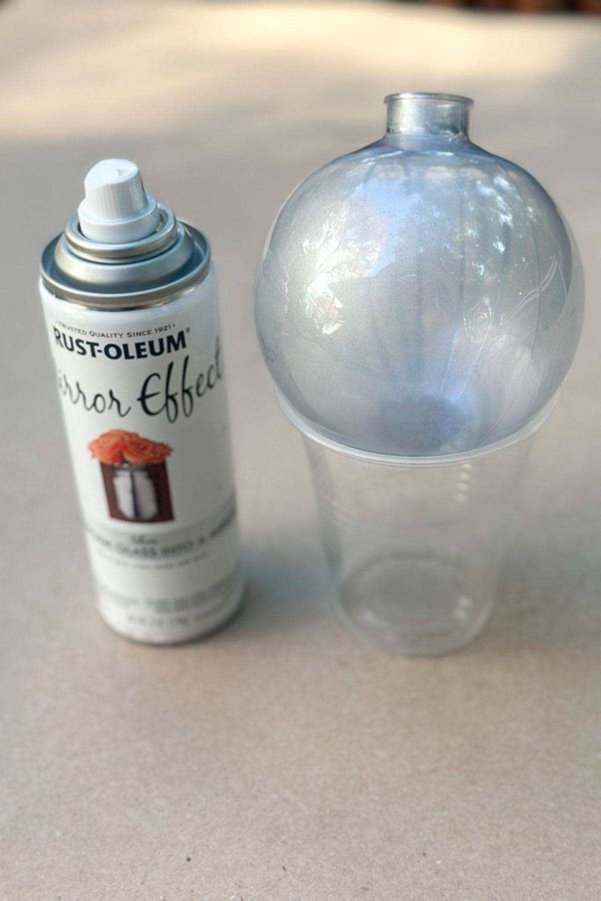 A can of Rust-Oleum Mirror Effect spray paint is next to a transparent plastic ornament placed on a clear plastic cup. The ornament's surface reflects light, indicating it has been partially sprayed to create a mirrored effect.